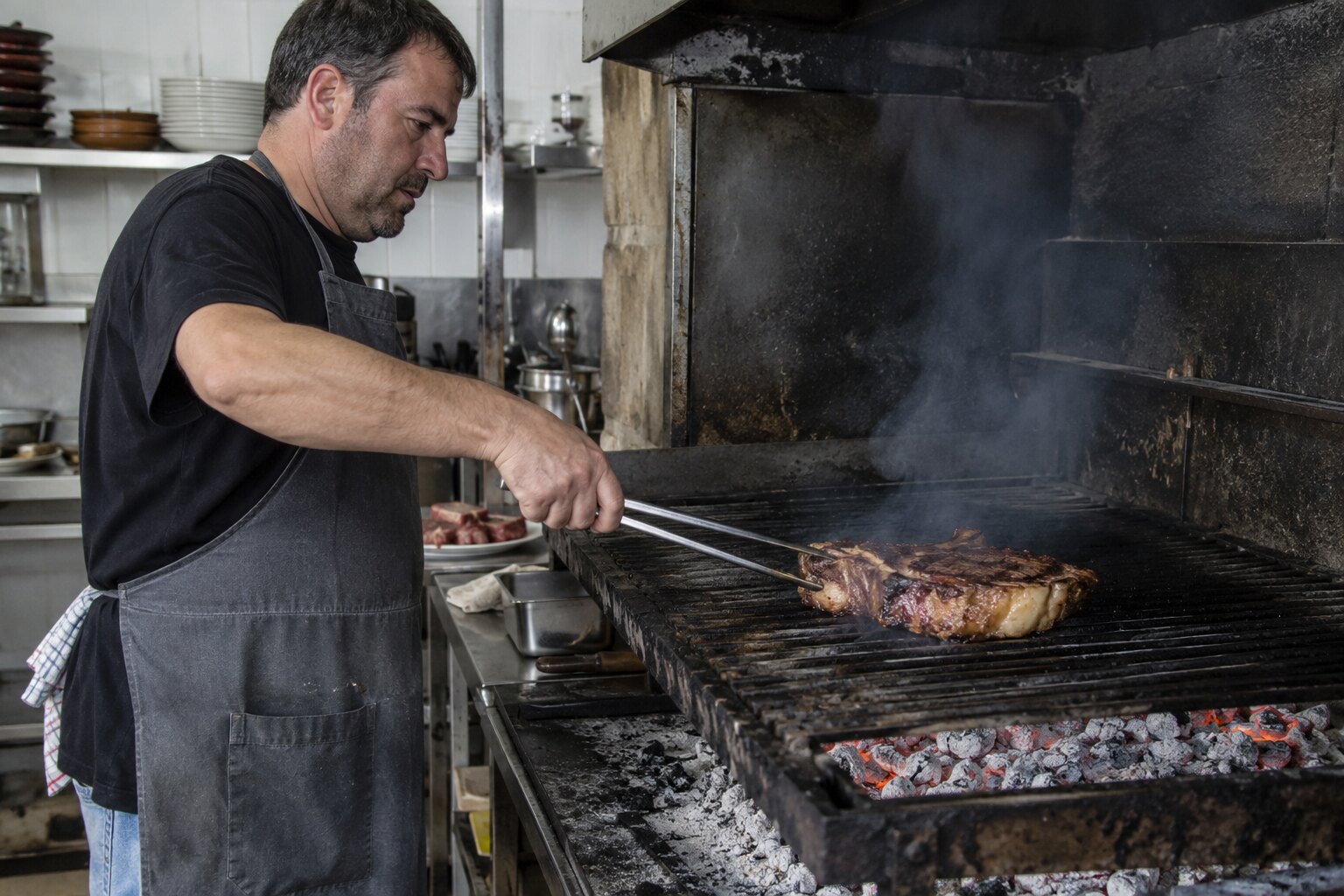 A cook working the grill at G cafe Bilbao, flames licking a thick Basque chuletón